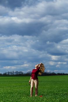 Beautiful young blonde woman stands on green grass in a park. Blue sky with clouds. The girl smiles and enjoys a good day.