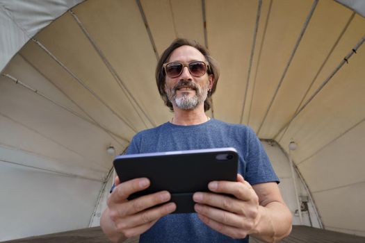 Sitting under tent covered concert venue middle aged grey haired freelancer man hold digital tablet in hands. Mature man talking or listening music use wireless earphones travel old town.