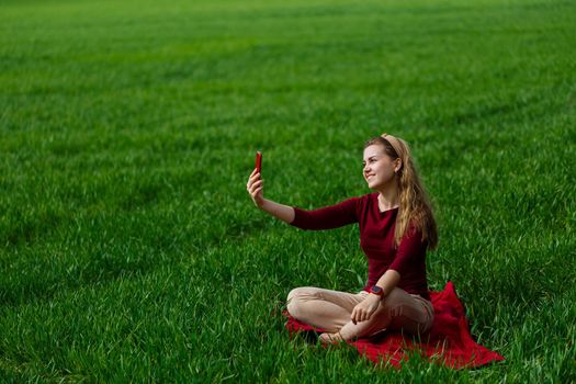 Beautiful woman with a phone in her hands sits on the grass. The girl looks at herself in the camera of the phone and takes selfie photos. She smiles and enjoys a warm day. Concept photo on smartphone