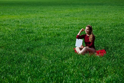 Young successful woman is sitting on green grass with a laptop in her hands. Rest after a good working day. Work on the nature. Student girl working in a secluded place. New business ideas