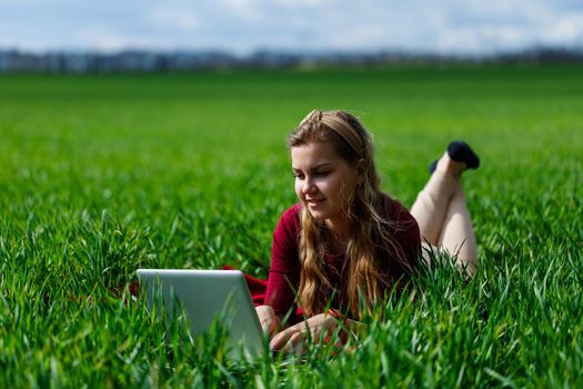 Beautiful young blonde woman is lying on the green grass in the park with a laptop and working. Blue sky with clouds. The girl smiles and enjoys a good day. Work on the nature on a sunny day.