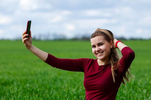 Beautiful woman with a phone in her hands sits on the grass. The girl looks at herself in the camera of the phone and takes selfie photos. She smiles and enjoys a warm day. Concept photo on smartphone