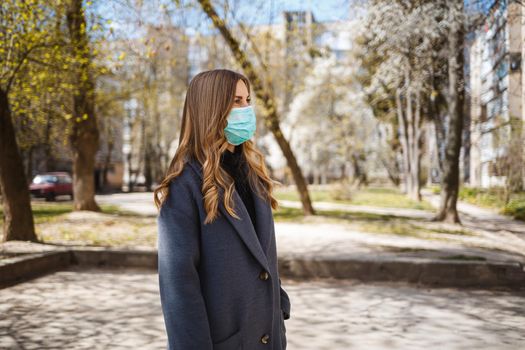 Girl, young woman in a protective sterile medical mask on her face, looking at the camera outdoors, in the spring garden. Air pollution, virus, pandemic coronavirus concept.