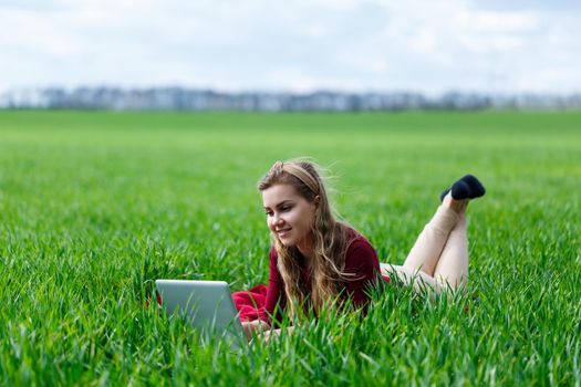 Beautiful young blonde woman is lying on the green grass in the park with a laptop and working. Blue sky with clouds. The girl smiles and enjoys a good day. Work on the nature on a sunny day.