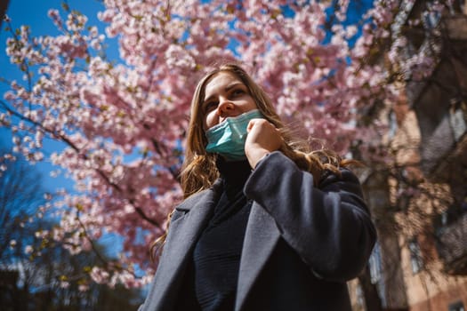 Girl, young woman in a protective sterile medical mask on her face, looking at the camera outdoors, in the spring garden. Air pollution, virus, pandemic coronavirus concept.