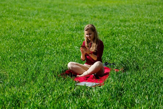 Young beautiful woman student with a phone in her hands sitting on the grass. Girl takes selfies and takes selfie pictures. She smiles and enjoys a warm day. Concept photo on smartphone