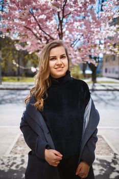 A young slender female model with long wavy hair and, dressed in a gray coat, sneakers, stands on the street near a flowering shrub with beautiful pink flowers in the background and poses.