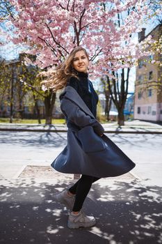 Young slender female model with long wavy hair and, dressed in a gray coat, sneakers, spinning on the street. Spring blossom trees woman girl laughs and runs to rejoice