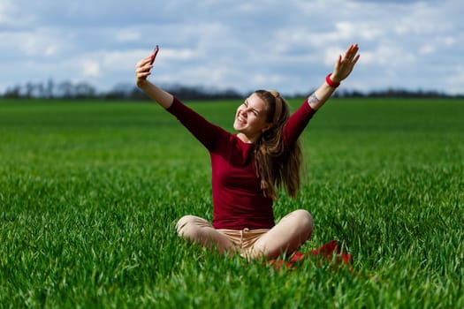 Young beautiful woman student with a phone in her hands sitting on the grass. Girl takes selfies and takes selfie pictures. She smiles and enjoys a warm day. Concept photo on smartphone