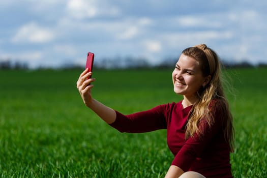 Beautiful woman with a phone in her hands sits on the grass. The girl looks at herself in the camera of the phone and takes selfie photos. She smiles and enjoys a warm day. Concept photo on smartphone