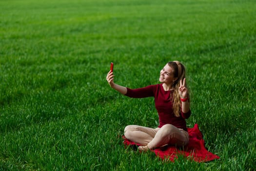 Beautiful woman with a phone in her hands sits on the grass. The girl looks at herself in the camera of the phone and takes selfie photos. She smiles and enjoys a warm day. Concept photo on smartphone