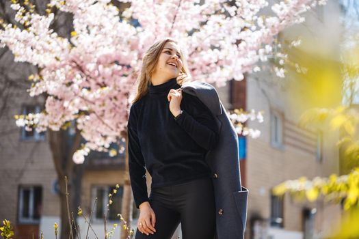 Sakura branches with flowers on a tree on the city streets. Happy woman girl in a gray palette walks along an alley with blooming sakura. Gorgeous fancy girl outdoors. Sakura tree blooming.