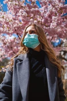 Girl, young woman in a protective sterile medical mask on her face, looking at the camera outdoors, in the spring garden. Air pollution, virus, pandemic coronavirus concept.