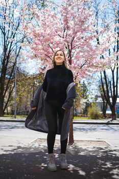 Young slender female model with long wavy hair and, dressed in a gray coat, sneakers, spinning on the street. Spring blossom trees woman girl laughs and runs to rejoice