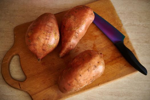 Top view of washed organic sweet potato tubers, batata, yam and kitchen knife on a wooden cutting board on kitchen countertop. Healthy eating. Raw vegan food. Food background. Root vegetable