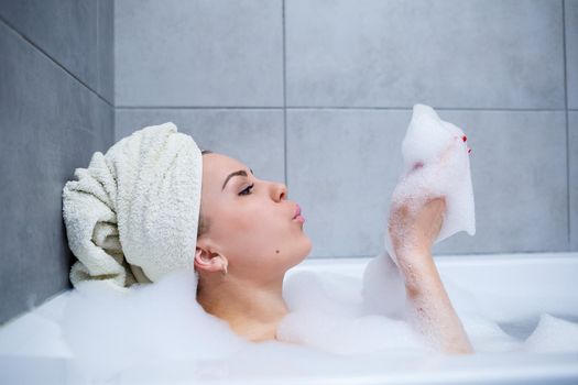 Girl woman with a white towel on her head lies in the bathroom in a white bath. In the hands of a lot of muddy soap. Full whirlpool foam. Relax after a hard day. Spa relaxing treatment
