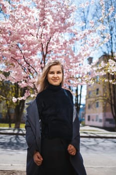 A young slender female model with long wavy hair and, dressed in a gray coat, sneakers, stands on the street near a flowering shrub with beautiful pink flowers in the background and poses.