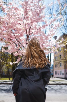 Young slender female model with long wavy hair and, dressed in a gray coat, sneakers, spinning on the street. Spring blossom trees woman girl laughs and runs to rejoice