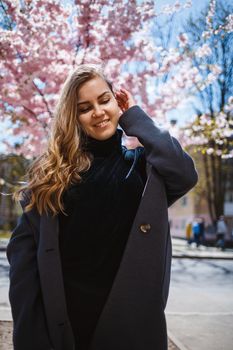 A young slender female model with long wavy hair and, dressed in a gray coat, sneakers, stands on the street near a flowering shrub with beautiful pink flowers in the background and poses.