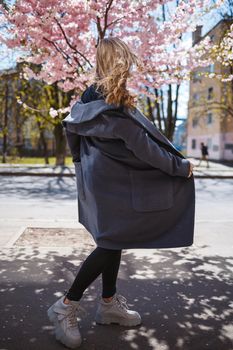 Young slender female model with long wavy hair and, dressed in a gray coat, sneakers, spinning on the street. Spring blossom trees woman girl laughs and runs to rejoice