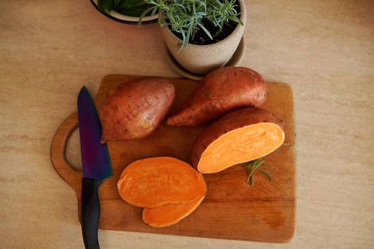 Overhead view of a whole and sliced tuber of sweet potato, batata, a kitchen knife on a wooden cutting board and cultivated rosemary in ceramic pot for meal seasoning on a beige kitchen countertop