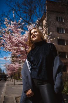 Sakura branches with flowers on a tree on the city streets. Happy woman girl in a gray palette walks along an alley with blooming sakura. Gorgeous fancy girl outdoors. Sakura tree blooming.