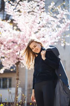 Sakura branches with flowers on a tree on the city streets. Happy woman girl in a gray palette walks along an alley with blooming sakura. Gorgeous fancy girl outdoors. Sakura tree blooming.