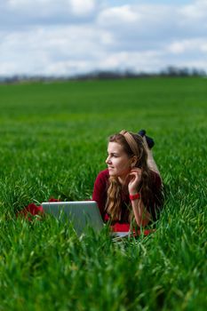 Beautiful young blonde woman is lying on the green grass in the park with a laptop and working. Blue sky with clouds. The girl smiles and enjoys a good day. Work on the nature on a sunny day.