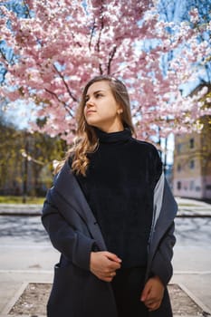 A young slender female model with long wavy hair and, dressed in a gray coat, sneakers, stands on the street near a flowering shrub with beautiful pink flowers in the background and poses.