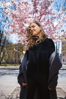 Young slender female model with long wavy hair and, dressed in a gray coat, sneakers, spinning on the street. Spring blossom trees woman girl laughs and runs to rejoice