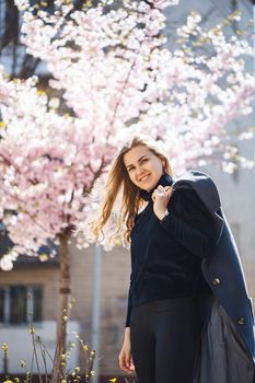 Sakura branches with flowers on a tree on the city streets. Happy woman girl in a gray palette walks along an alley with blooming sakura. Gorgeous fancy girl outdoors. Sakura tree blooming.