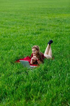 Beautiful young blonde woman is lying on the green grass in the park with a laptop and working. Blue sky with clouds. The girl smiles and enjoys a good day. Work on the nature on a sunny day.