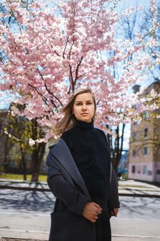 A young slender female model with long wavy hair and, dressed in a gray coat, sneakers, stands on the street near a flowering shrub with beautiful pink flowers in the background and poses.