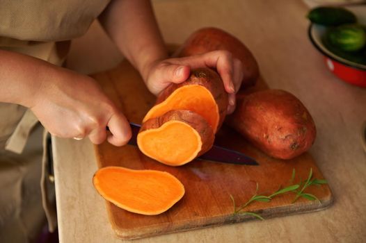 Selective focus on female hands slicing organic raw tuber of a whole sweet potato on a wooden cutting board. Batata. Yam.