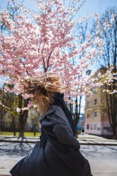 Young slender female model with long wavy hair and, dressed in a gray coat, sneakers, spinning on the street. Spring blossom trees woman girl laughs and runs to rejoice. Selective focus