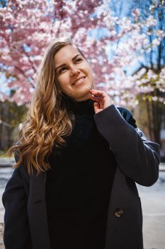 A young slender female model with long wavy hair and, dressed in a gray coat, sneakers, stands on the street near a flowering shrub with beautiful pink flowers in the background and poses.