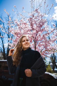 Sakura branches with flowers on a tree on the city streets. Stylish woman girl stands in the street with blooming sakura. Modern fashionable girl outdoors. Sakura tree blossoms.