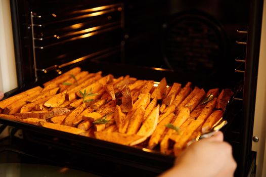 Selective focus on sliced wedges of sweet potatoes, seasoned with spices and culinary herbs, sprinkled with olive oil, on a baking sheet while placing in into an oven. Cooking fried, roasted batata