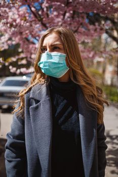 Girl, young woman in a protective sterile medical mask on her face, looking at the camera outdoors, in the spring garden. Air pollution, virus, pandemic coronavirus concept.