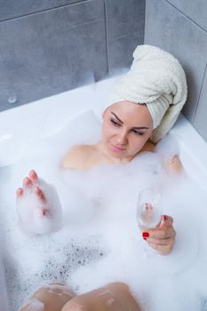 Girl woman with a white towel on her head lies with a glass of champagne in a white bathtub. In her hands a lot of pee soap. Full whirlpool foam. Relax after a hard day. Spa relaxing procedure