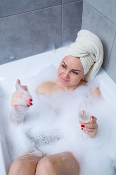 Girl woman with a white towel on her head lies with a glass of champagne in a white bathtub. In her hands a lot of pee soap. Full whirlpool foam. Relax after a hard day. Spa relaxing procedure
