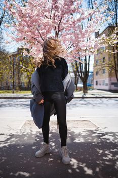 Young slender female model with long wavy hair and, dressed in a gray coat, sneakers, spinning on the street. Spring blossom trees woman girl laughs and runs to rejoice