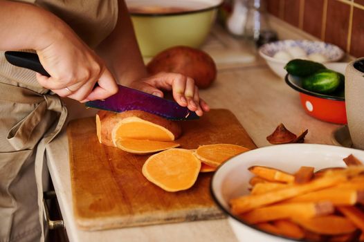 Close-up housewife in beige chef's apron, preparing healthy vegan food, cutting a whole tuber of sweet potato into slices on a wooden chopping board, standing at a kitchen countertop. Selective focus