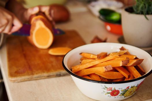 Selective focus on an enamel vintage bowl with wedges of sweet potato, against the blurred background of chef's hands using a kitchen knife, cutting whole tubers of organic batata on a wooden board