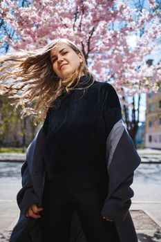 Young slender female model with long wavy hair and, dressed in a gray coat, sneakers, spinning on the street. Spring blossom trees woman girl laughs and runs to rejoice