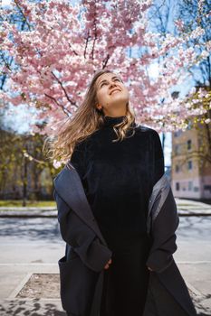 Young slender female model with long wavy hair and, dressed in a gray coat, sneakers, spinning on the street. Spring blossom trees woman girl laughs and runs to rejoice