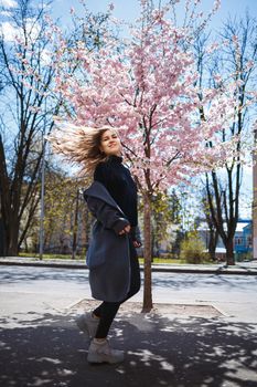 Young slender female model with long wavy hair and, dressed in a gray coat, sneakers, spinning on the street. Spring blossom trees woman girl laughs and runs to rejoice