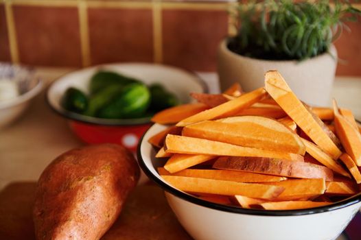 Selective focus on wedges of organic sweet potato in an enamel vintage bowl next to tubers of batata, on a wooden cutting board, against a blurred pot with rosemary in a kitchen countertop