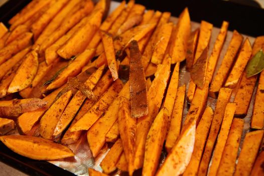 Healthy homemade sweet potato fries on a baking sheet. Delicious vegan meal - roasted batata wedges baked with rosemary leaves. Close-up. Delicious healthy vegan meal.