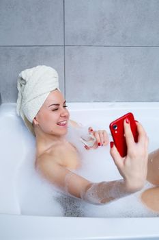 Girl woman with a towel on her head lies with a glass of champagne in a white bathtub and takes a selfie on the phone. In her hands a lot of drunk soap. Relax after a hard day. Spa relaxing procedure
